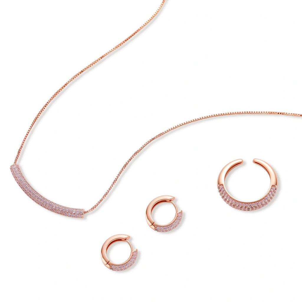 Rose gold pavé zirconia bar necklace, hoop earrings, and open ring set with pink stones on cream background.